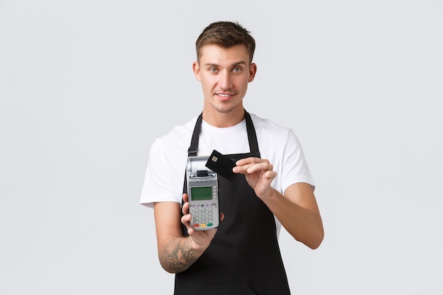 Commerce de détail, café et restaurant, concept d'employés. Beau serveur sympathique et poli donnant un terminal de point de vente, client payant un café avec une carte de crédit, souriant joyeusement, fond blanc