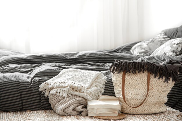 Composition avec grand sac en paille en osier, couvertures et livres sur fond de chambre.