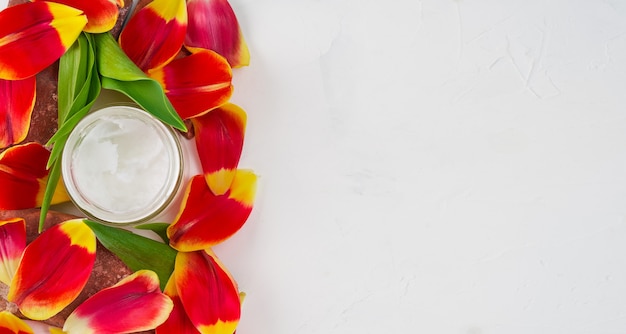 Composition avec de l'huile de noix de coco dans un pot sur blanc entouré de pétales de tulipes, vue de dessus avec espace copie