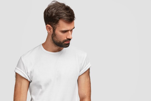 Concentré beau jeune homme concentré vers le bas, a le chaume et les cheveux foncés, porte un t-shirt blanc décontracté