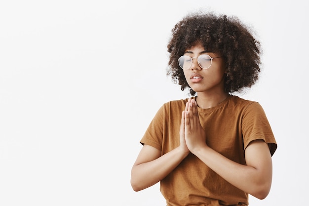 concentré détendu et calme attrayant jeune femme à la peau foncée dans des lunettes avec une coiffure afro debout à moitié tourné vers la gauche avec les yeux fermés geste namaste ou les mains en priant méditer