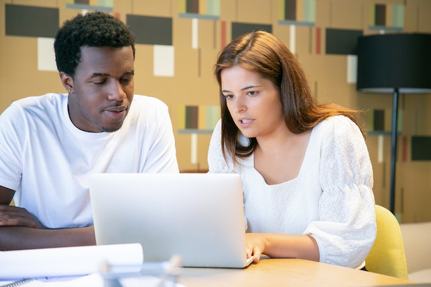 Concentré divers designers regardant ensemble la présentation du projet sur un ordinateur portable, assis à table et parlant, discutant du projet de conception