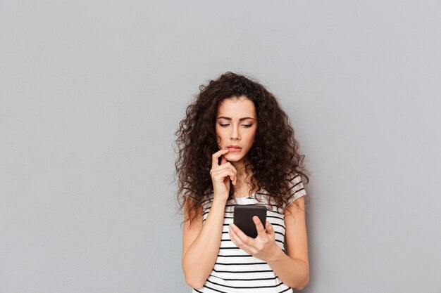 Concentré de jeune femme à l'aide de smartphone et de toucher ses lèvres frustrés ou recevoir de mauvaises nouvelles sur le mur gris