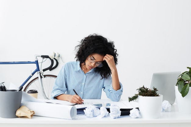 Concentré jeune femme à la peau sombre portant des lunettes et une chemise bleue assise sur son lieu de travail