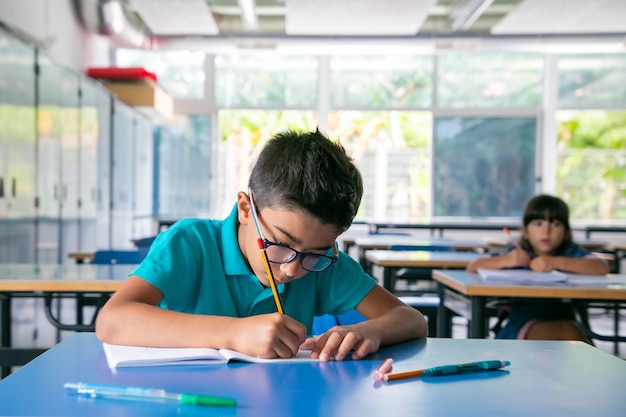 Concentré jeune garçon dans des verres assis au bureau et écrit dans un cahier en classe