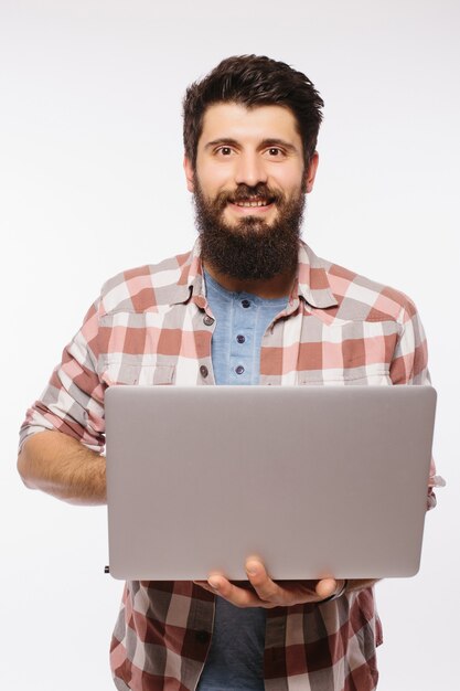 Concentré jeune homme barbu portant des lunettes habillé en chemise à l'aide d'un ordinateur portable isolé sur un mur blanc.