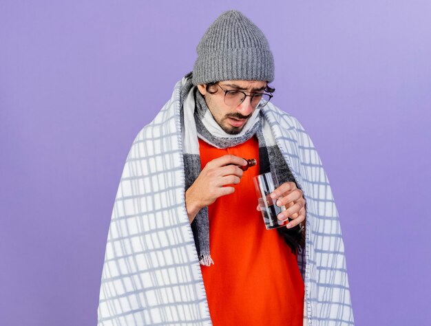 Concentré jeune homme malade de race blanche portant des lunettes chapeau d'hiver et écharpe enveloppé dans un plaid versant un médicament en verre dans un verre d'eau isolé sur un mur violet avec espace de copie