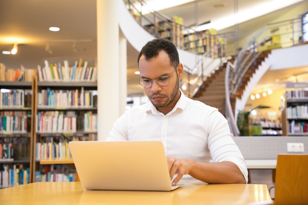 Concentré de jeune homme tapant sur un ordinateur portable à la bibliothèque publique