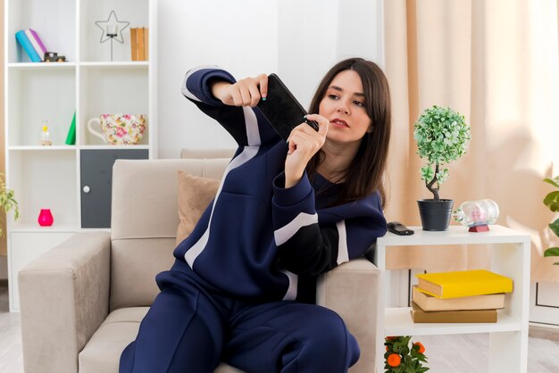 Concentré jeune jolie femme caucasienne assise sur un fauteuil dans un salon conçu à jouer au jeu sur téléphone