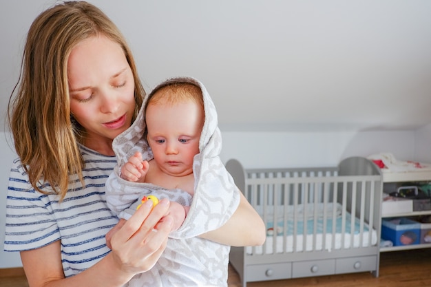 Photo gratuite concentré jeune maman tenant doux bébé sec enveloppé dans une serviette à capuchon après la douche, jouant avec un jouet de bain en caoutchouc. vue de face, copiez l'espace. concept de garde d'enfants ou de bain