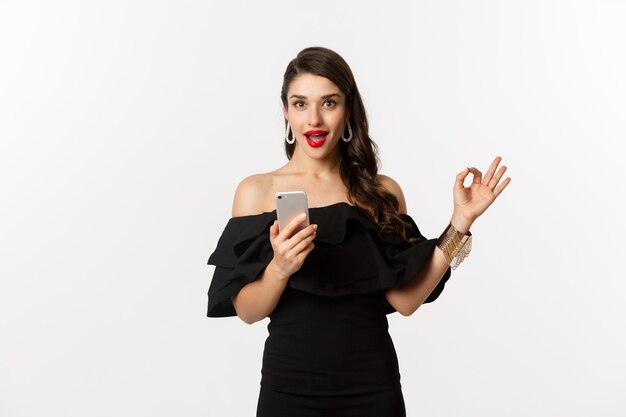 Concept d'achat en ligne. Jolie femme en robe noire à la mode, maquillage, montrant l'approbation de l'enregistrement et utilisant l'application pour téléphone portable, fond blanc