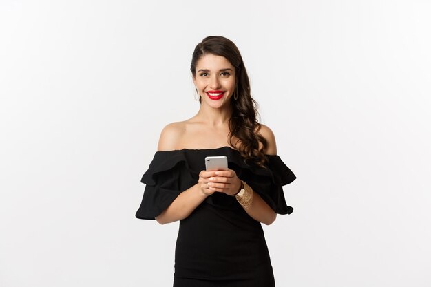 Concept d'achat en ligne. Jolie jeune femme en robe noire, lisant un message texte, utilisant un téléphone portable et souriant, debout sur fond blanc