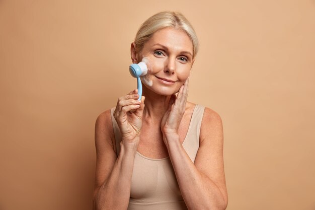 Concept d'âge et d'hygiène de beauté. Jolie femme blonde senior ridée utilise un masseur pour le visage et lave les joues avec de la mousse habillée en haut décontracté a une peau saine parfaite