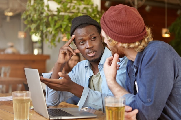 Concept d'amitié interraciale. Deux meilleurs amis portant des chapeaux assis à la table du café et parlant, discutant de plans, partageant des nouvelles, buvant de la bière et regardant un match de football sur un ordinateur latop générique