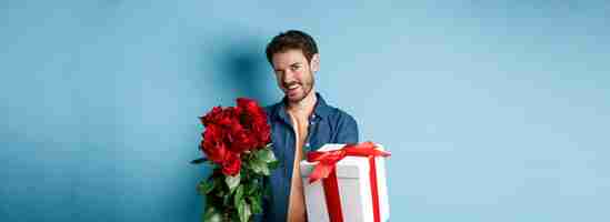Photo gratuite concept d'amour et de saint valentin mec heureux faisant un clin d'œil et souriant à la caméra présentant un cadeau et une bouque