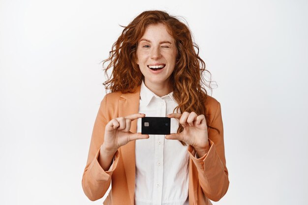 Concept d'argent et de finances Vendeuse rousse souriante montrant une carte de crédit et un clin d'œil debout en costume sur fond blanc