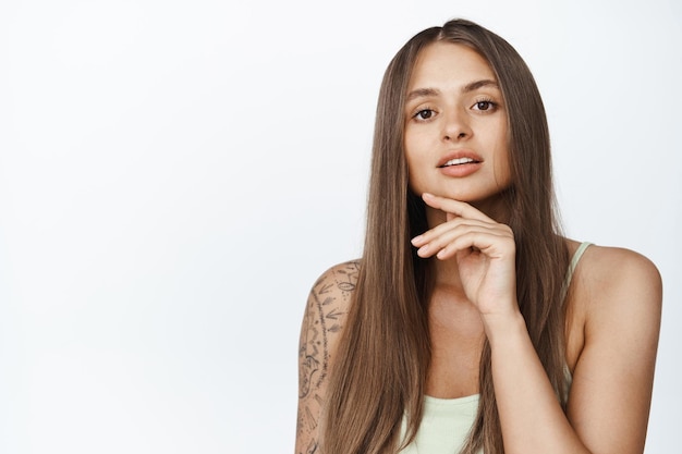 Concept de beauté et de maquillage pour femmes. Jeune femme élégante touche son visage, l'air pensif à la caméra, a une peau lisse et saine et de longs cheveux naturels, fond blanc