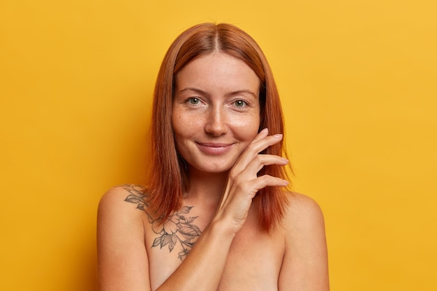 Concept de beauté naturelle, de pureté et de bien-être. Une femme rousse satisfaite touche doucement le visage, démontre sa peau lisse parfaite après la procédure de spa, se tient les épaules nues contre le mur jaune