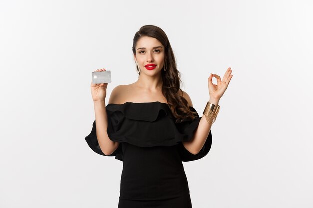 Concept de beauté et de shopping. Superbe femme en bijoux de luxe et robe noire, montrant un signe correct et une carte de crédit, debout sur fond blanc.