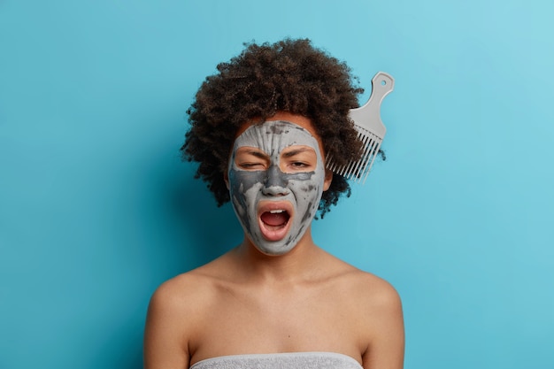 Concept de beauté de soins du corps. Belle femme à la peau foncée applique un masque facial a un peigne coincé dans les cheveux bouclés bâille