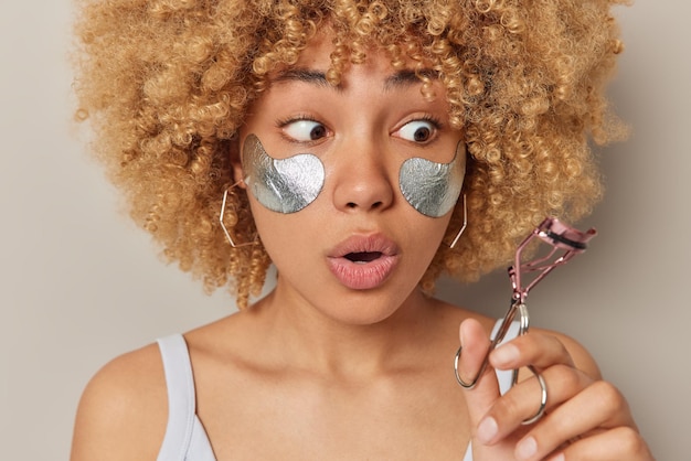 Photo gratuite concept de beauté et de soins de la peau une femme aux cheveux bouclés choquée applique des patchs argentés de beauté sous les yeux regarde avec étonnement les cils le recourbe-cils ne sait pas comment utiliser cet instrument pose à l'intérieur