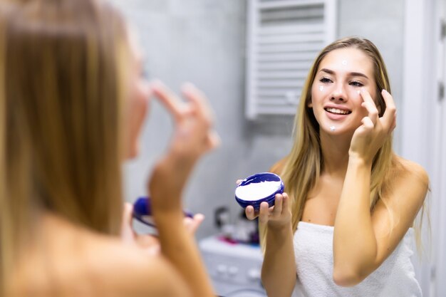 Concept de beauté, de soins de la peau et de personnes - gros plan sur une jeune femme appliquant de la crème sur le visage et regardant un miroir dans la salle de bain de la maison