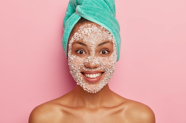 Photo gratuite concept de beauté et de soins de santé. la femme à la peau foncée satisfaite applique un masque facial exfoliant blanc, se soucie de la pureté et de la douceur de son teint, a une serviette turquoise sur la tête