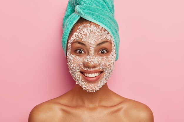 Concept de beauté et de soins de santé. La femme à la peau foncée satisfaite applique un masque facial exfoliant blanc, se soucie de la pureté et de la douceur de son teint, a une serviette turquoise sur la tête