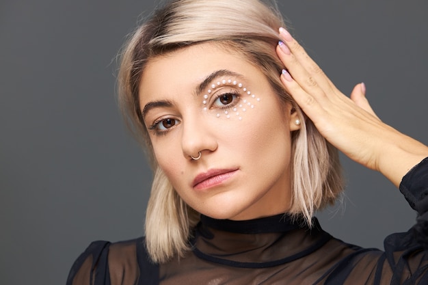 Concept de beauté, style et féminité. Superbe femme européenne sensuelle avec une coiffure bob blonde démontrant son maquillage élégant, cristaux blancs autour des yeux