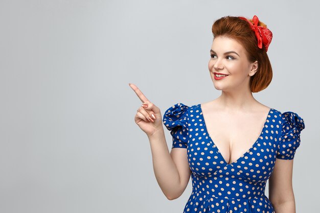 Concept de beauté, style, mode et élégance. Jolie fille habillée en pin up style vintage montrant l'espace de copie pour votre information, pointant le doigt avant sur le mur du studio vide et souriant largement