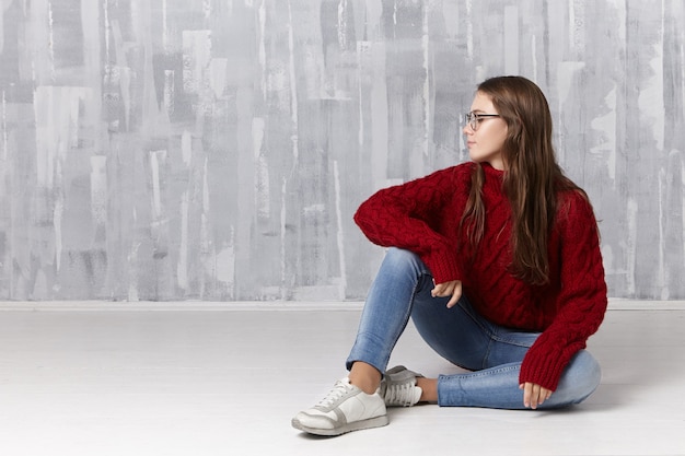 Concept de beauté, style, mode, jeunesse, personnes et style de vie. Belle charmante adolescente aux longs cheveux lâches assis sur le sol, portant des lunettes, un pull à col roulé, des jeans et des baskets