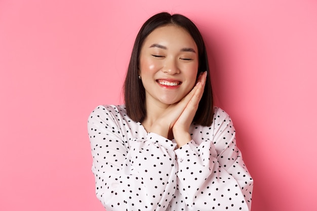 Concept de beauté et de style de vie. Gros plan d'une femme asiatique belle et rêveuse dormant sur ses mains, fermer les yeux et souriant, rêvasser sur fond rose.