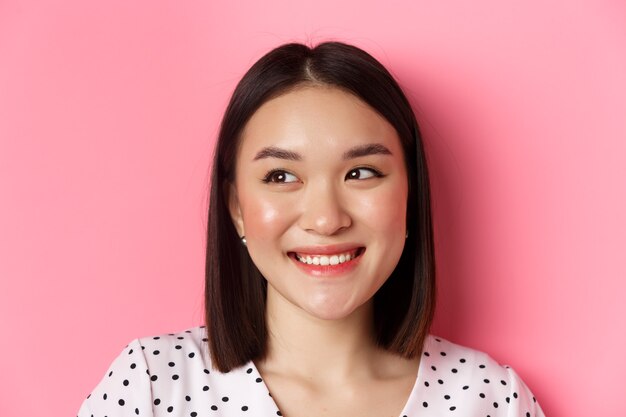 Concept de beauté et de style de vie. Portrait d'une jolie femme asiatique rêveuse regardant à gauche l'espace de copie, souriante heureuse, debout sur fond rose