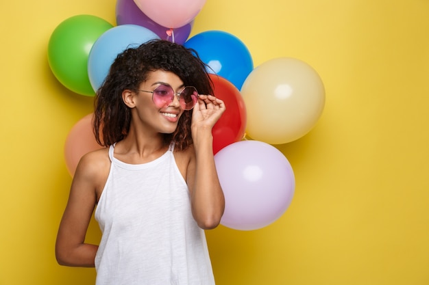 Concept de célébration - Gros plan Portrait heureux jeune femme africaine belle en t-shirt noir souriant avec un ballon de partie coloré. Fond d&#39;écran en pastel jaune pastel.