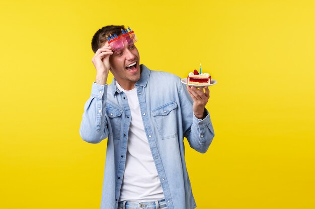 Concept de célébration, de vacances et d'émotions de personnes. Heureux joyeux anniversaire beau mec, lunettes de soleil au décollage et émerveillé par un délicieux gâteau d'anniversaire avec une bougie, fond jaune.