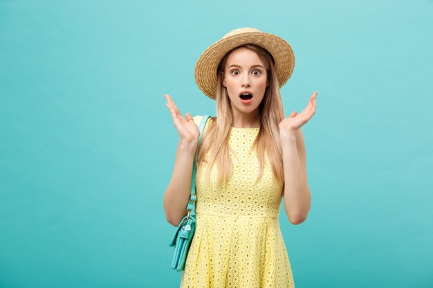 Concept de choc ou de bonne nouvelle : Jeune fille blonde élancée aux cheveux longs dans un chapeau se dresse sur un fond bleu isolé. Elle ouvrit la bouche de surprise.