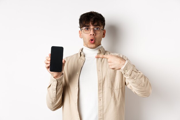 Concept de commerce électronique. Portrait de jeune homme pointant sur l'écran du téléphone portable, montrant une publicité en ligne, debout sur fond blanc