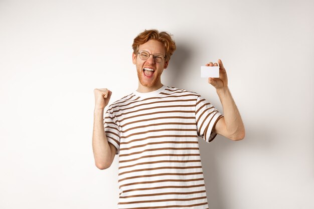 Concept commercial et financier. Jeune homme remportant un prix bancaire, montrant une carte de crédit en plastique et faisant une pompe à poing, criant de joie et de satisfaction, fond blanc