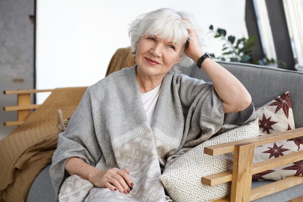 Concept de confort, design d'intérieur, loisirs, mode de vie et personnes âgées. Attractive élégante femme mûre retraité avec des rides et des cheveux gris reposant sur un canapé dans sa maison de campagne, souriant