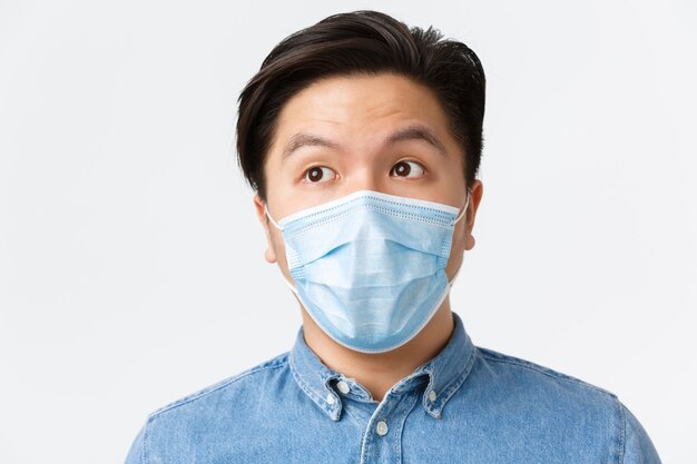 Concept de coronavirus, de distanciation sociale et de style de vie. Gros plan d'un entrepreneur asiatique réfléchi portant un masque médical, regardant le coin supérieur gauche en train de réfléchir, faisant son choix, fond blanc.