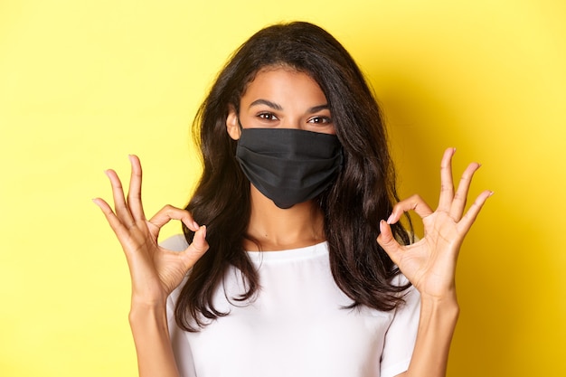 Concept de coronavirus, pandémie et mode de vie. Gros plan d'une femme afro-américaine confiante et souriante portant un masque noir, montrant des signes d'approbation, comme quelque chose de bien, fond jaune.