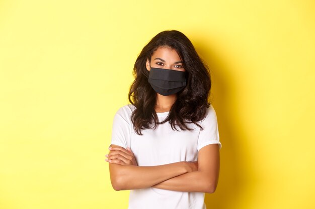 Concept de coronavirus, pandémie et mode de vie. Portrait d'une jeune femme afro-américaine au masque noir, souriante et semblant confiante, les mains croisées sur la poitrine, fond jaune.