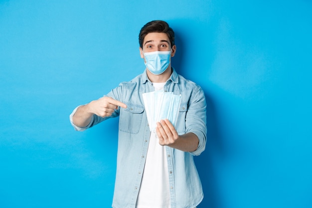 Concept de coronavirus, quarantaine et distanciation sociale. Jeune homme pointant sur des masques médicaux, empêchant les mesures de covid-19, debout sur fond bleu