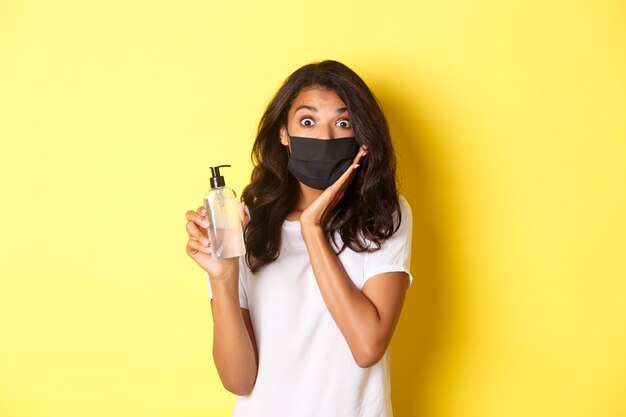 Concept de covid-19, distanciation sociale et mode de vie. Image d'une fille afro-américaine impressionnée en masque facial, montrant un bon désinfectant pour les mains, recommandant un produit, fond jaune