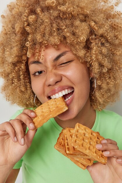 Concept de la dent sucrée Gros plan d'une jeune femme aux cheveux bouclés mord des gaufres avec appétit cligne de l'œil vêtu de vêtements décontractés rompt le régime alimentaire Modèle féminin avec des pâtisseries fraîches Nourriture délicieuse