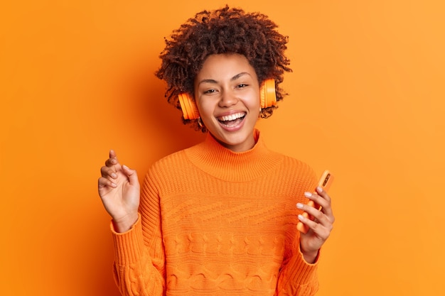 Concept de divertissement et de loisirs de personnes. Enthousiaste jeune femme afro-américaine aux cheveux bouclés détient smartphone moderne écoute de la musique via des écouteurs stéréo pose sur fond orange vif