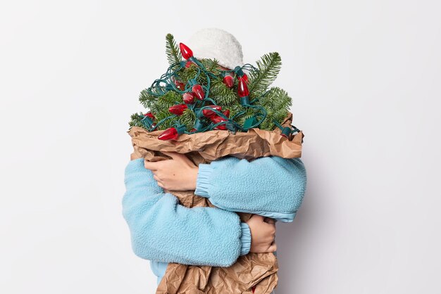 Photo gratuite concept du nouvel an et de noël. une femme méconnaissable se cache derrière un grand bouquet de sapins décoré de guirlandes attend pour les vacances à venir vêtues de poses de vêtements d'extérieur sur fond blanc.