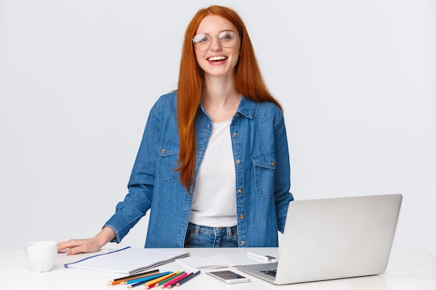 Concept d'éducation, de travail et d'indépendant. Nomade numérique féminine rousse charismatique attrayante, designer travaillant à distance, créant un projet de conception, debout près d'une table avec un ordinateur portable, des crayons de couleur.