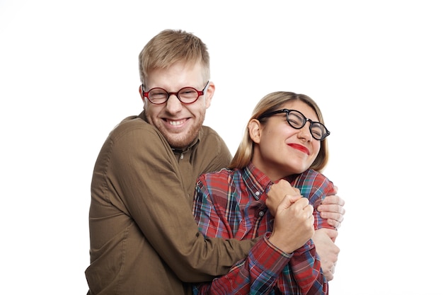 Concept d'émotions humaines positives, de bonheur et de joie. Tir horizontal de joyeux jeune couple à lunettes s'amuser: barbu avec grimace tenant une femme souriante serrée