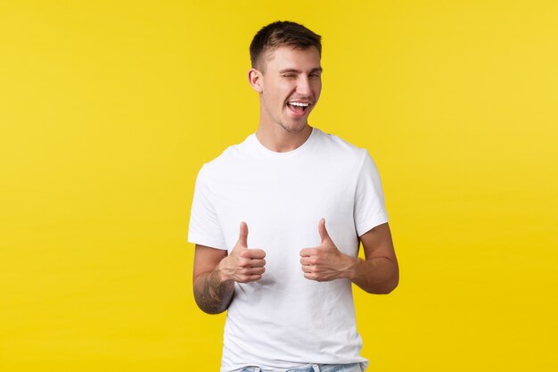 Concept d'émotions de mode de vie, d'été et de personnes. Un client masculin satisfait et effronté en t-shirt blanc basique, un clin d'œil et un sourire encouragent à essayer, recommandent le produit avec le pouce levé, fond jaune.